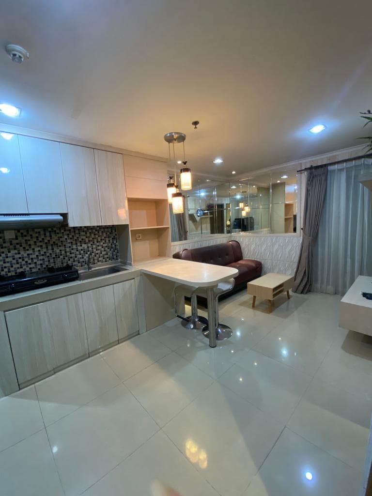Dapur dengan kompor gas, sink, dan kitchen set menyatu dengan ruang keluarga berisi sofa dan meja bar di unit 1BR Green Central City